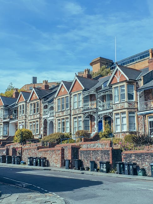 A row of Victorian-style terraced houses situated on a slightly elevated street in West Hampstead, featuring detailed ironwork balconies, decorative gables, large bay windows, and brick facades. The houses are built with a combination of red brick and light-colored stone detailing, with some having small front gardens enclosed by low brick walls and shrubbery. The street below has a row of black wheelie bins and a paved pavement, with a clear blue sky overhead, indicating a bright, sunny day. The image captures the exterior of the properties, emphasizing the architectural features typical of the area, which are relevant in the context of home relocation or furniture transport as handled by [COMPANY_NAME]. This scene underscores the importance of careful planning during house removals in West Hampstead, including loading and transport processes for residential furniture and belongings.