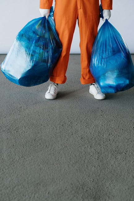 A person standing indoors on a grey carpeted floor, dressed in orange work trousers and white sneakers, holding two large blue plastic rubbish bags filled with waste. The individual’s hands, covered w