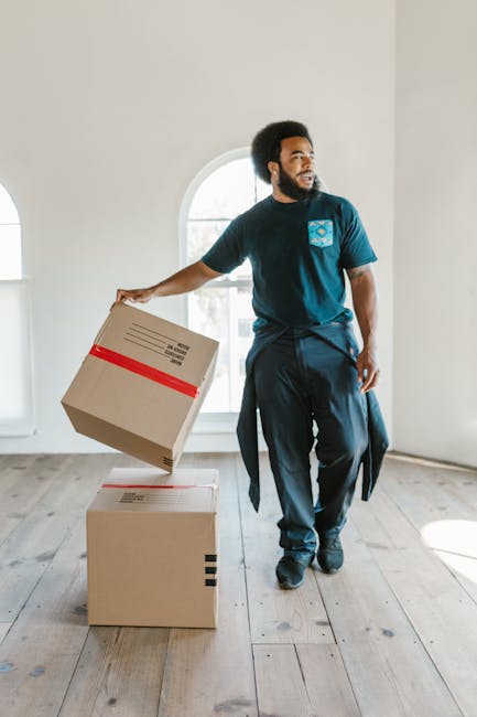 A man with dark curly hair and a beard, dressed in a blue T-shirt and dark trousers, is lifting a cardboard box wrapped with red tape in a bright, spacious room with wooden flooring and white walls. Two other cardboard boxes, one with black tape, are placed on the floor nearby. The room features large arched windows allowing natural light to fill the space, and the scene appears to depict a home relocation or furniture transport process. This natural interior setting illustrates packing and moving activities typical of house removals services, such as those offered by [COMPANY_NAME], highlighting the careful handling of packed belongings during a professional house or furniture move.