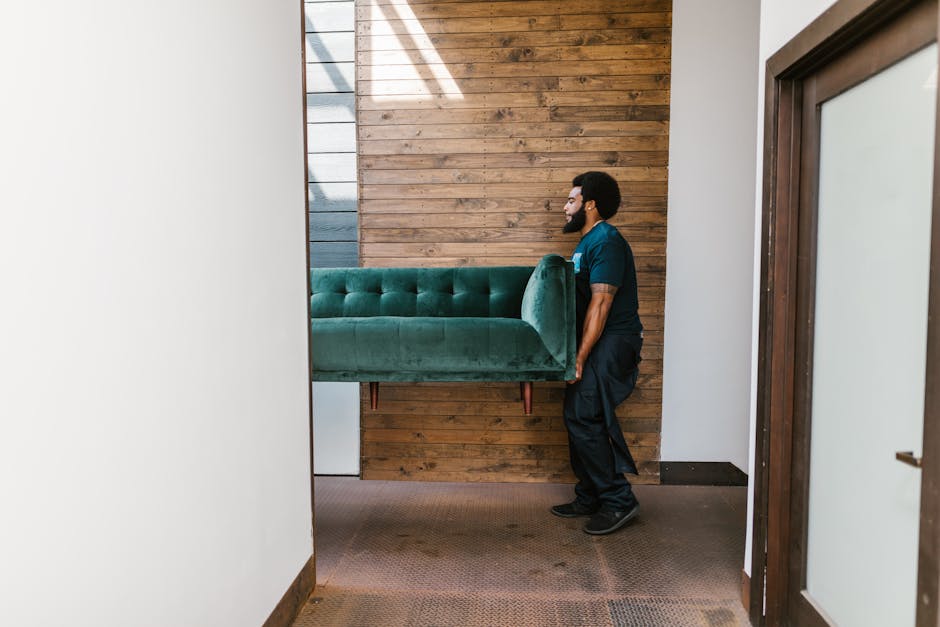 A professional furniture mover is inside a residential property, carefully lifting a green velvet sofa with wooden legs. The mover, dressed in dark work attire, is positioned to the right of the image, supporting the sofa from beneath with both hands. The room has a wooden accent wall behind them, composed of horizontal planks, with natural light streaming in from the left side, casting shadows across the space. The interior features white walls, and the sofa is being prepared for transport as part of a home relocation process. The entrance to the room is framed by a door on the right, and the floor is carpeted in a neutral tone. This image illustrates a typical packing and moving activity involved in furniture transport, highlighting the careful handling required during the loading process for house removals, as is often managed by companies like [COMPANY_NAME]. The scene captures the logistical aspect of residential furniture moving within a domestic setting, emphasizing the importance of proper lifting techniques and equipment such as trolleys or straps employed in professional removals.