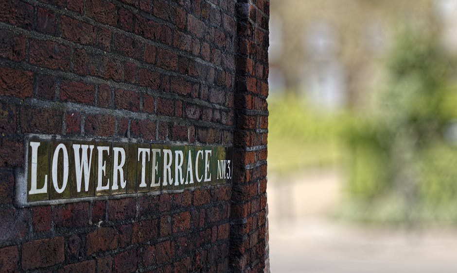 Close-up view of a brick wall featuring a street sign reading 'LOWER TERRACE NW3' in white lettering on a rectangular, slightly weathered metal plaque, attached to the bricks. To the right, an open doorway or entrance is visible, leading into a property where packing materials such as cardboard boxes and plastic-wrapped furniture items are being moved. Inside the property, there are cardboard boxes stacked near the entrance, with some wrapped furniture pieces laid out on the floor, indicating active packing and furniture transport preparations. A moving trolley with a blue strap and a moving blanket draped over furniture can be seen, suggesting the process of loading items for a home relocation. Natural light illuminates the scene, highlighting the textures of the bricks, the packaging materials, and the furniture. The image is associated with professional removals services, subtly emphasizing house removals and logistical details involved in packing and moving operations, as provided by [COMPANY_NAME], supporting efficient moving logistics for residents in West Hampstead.