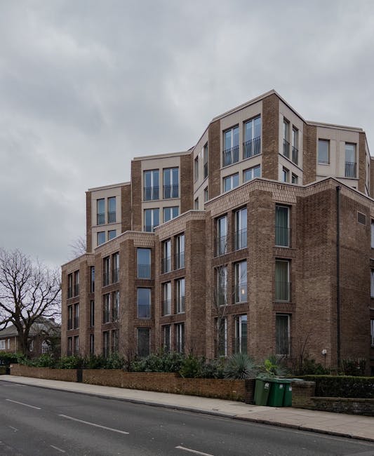 A multi-storey residential building made of brown and beige brick with large rectangular windows, situated along Maygrove Road near West Hampstead Thameslink station. In the foreground, the street features a pavement with two green waste bins and some small bushes and trees along the edge of the property. The building has a modern architectural style with multiple levels, some sections with protruding balconies and sectioned windows, and is under overcast sky with grey clouds. The scene captures the exterior of the property, highlighting its height and urban setting, indicative of a typical home relocation or furniture transport environment nearby provided by [COMPANY_NAME], engaging in professional removals and moving services.