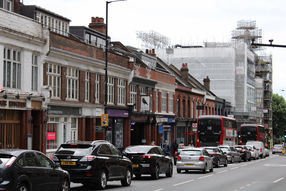 An elevated view of Maygrove Road near West Hampstead Thameslink station showing a single-lane road with parked cars along the curb and moving vehicles including vans and smaller cars, bordered by trees with green foliage on both sides. On the left, multi-storey brick buildings with flat roofs and commercial signage are visible, while on the right, there is a small, white, single-story building with a pitched roof, bicycle racks, and pedestrians walking along the sidewalks. In the background, residential and commercial buildings extend into a tree-lined street, highlighting typical urban surroundings for home relocation and furniture transport services; this scene captures a typical busy street environment where a removal company like [COMPANY_NAME] might load household items into a van for moving within or outside West Hampstead, involving parking, street navigation, and coordination with local infrastructure.