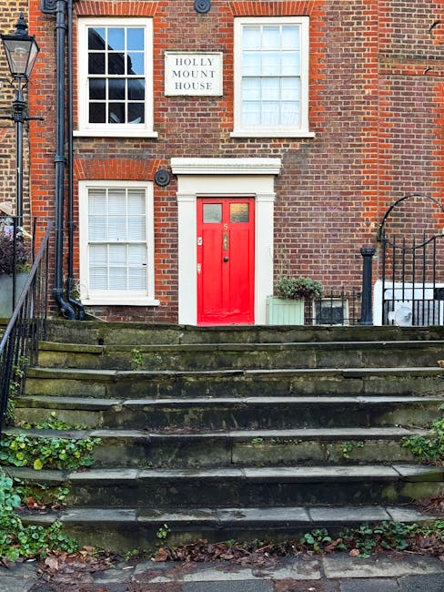 The image shows the front exterior of a traditional brick house with white-framed sash windows on the upper and lower floors. A prominent red front door is set within a white surround, accessible via a short flight of weathered stone steps with patches of moss and grass growing between them. On the left side of the entrance, there is a white sign reading 'Holly Mount House' mounted on the brick wall. To the right of the door, a small green plant in a rectangular planter sits on the pavement, next to a black metal gate that leads into the property. The setting includes an adjacent street lamp and a black metal railing lining the staircase. This exterior scene illustrates a typical Victorian terrace property in West Hampstead, providing context for house removals and furniture transport activities by [COMPANY_NAME], with visible elements relevant to packing and moving logistics including the staircase and the entrance area prepared for a relocation project.
