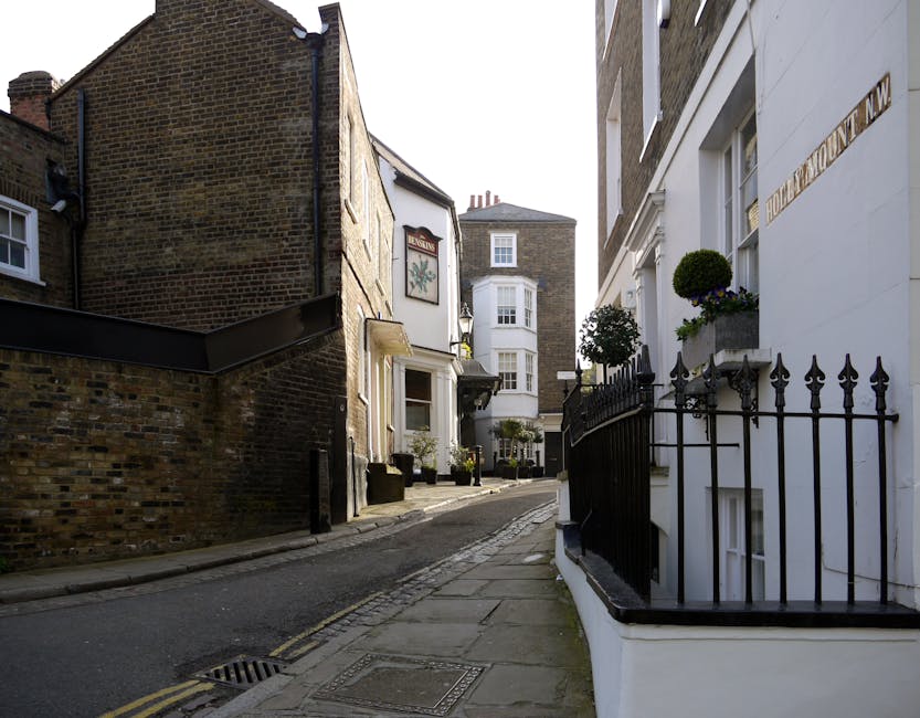 The image depicts a narrow residential street in West Hampstead with buildings on either side. On the right, there is a white building with a black wrought iron fence and potted plants on the windowsills, highlighting a Victorian-style terrace. The building on the left is constructed with exposed brickwork, featuring a small window and a slightly recessed doorway. The pavement is made of stone slabs, with a narrow, sloped road running through the centre of the scene. Some artistic potted plants and small trees are visible outside the buildings, and the street appears to be part of a quiet, historic neighbourhood. Given the context of house removals and the narrow access, this setting exemplifies the logistical challenges associated with furniture transport and home relocation in tight urban environments. This scene may be used by [COMPANY_NAME] in illustrating the process of planning for moving services through confined city streets, emphasizing careful loading and transport procedures involved in removals within West Hampstead.