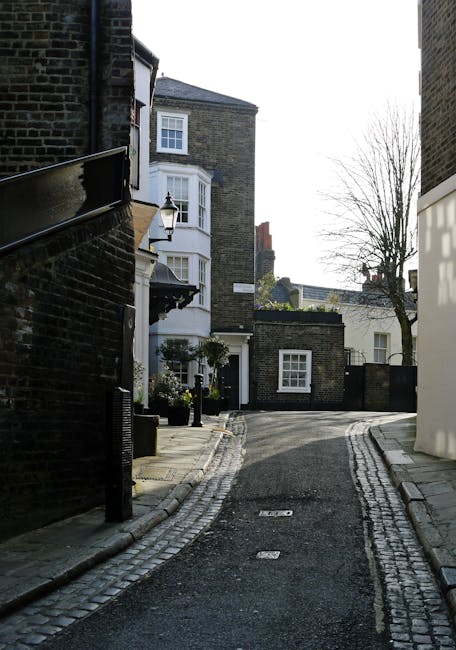 The image shows the front exterior of a traditional brick house with white-framed sash windows on the upper and lower floors. A prominent red front door is set within a white surround, accessible via a short flight of weathered stone steps with patches of moss and grass growing between them. On the left side of the entrance, there is a white sign reading 'Holly Mount House' mounted on the brick wall. To the right of the door, a small green plant in a rectangular planter sits on the pavement, next to a black metal gate that leads into the property. The setting includes an adjacent street lamp and a black metal railing lining the staircase. This exterior scene illustrates a typical Victorian terrace property in West Hampstead, providing context for house removals and furniture transport activities by [COMPANY_NAME], with visible elements relevant to packing and moving logistics including the staircase and the entrance area prepared for a relocation project.