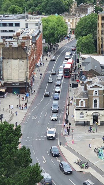 An aerial view of a residential street in West Hampstead, NW6, showing multiple vehicles including cars and buses traveling along a curved, one-way road. On the left side, there are pedestrians walking on the pavement, some waiting at crosswalks, and storefronts with signage, indicating local shops and businesses. On the right side, there are parked bicycles near a small building with classical architectural details, and a tree-lined sidewalk with pedestrians and cycling activity. Tall, leafy trees line both sides of the street, providing greenery amidst the urban environment. The scene is well-lit, suggesting daytime, and the overall setting depicts a typical busy neighborhood suitable for house removals and furniture transport, with logistics related to packing, loading, and ensuring smooth relocation processes. This image is relevant to [COMPANY_NAME]'s domestic moving services, showcasing the typical urban environment encountered during home relocations in NW6, West Hampstead.