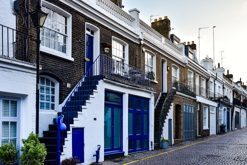 Exterior view of a row of terraced houses in West Hampstead with traditional brick facades, white window frames, and small balconies with black or blue railings. A house with a white and blue ground-floor entrance has a staircase with a blue railing leading to a blue door on the first floor. The cobblestone street is lined with potted plants and small bushes, and a large window with white shutters is visible on the lower level. The scene depicts daytime with natural light illuminating the building facades. This setting relates to house removals and furniture transport in narrow staircases, as evidenced by the staircase and available space for moving activities, supported by professional removals services like those from [COMPANY_NAME].