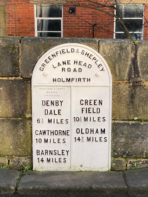 A white, vintage-style directional road sign mounted on a stone wall outside a brick building with two windows and a metal handrail, displaying distances to various locations, including Denby Dale, Cawthorne, Barnsley, Oldham, and Greenfield. The sign has multiple sections with black lettering, headings such as 'Greenfield & Shepley Lane Head Road,' and distances measured in miles, with some fractions like 6 1/2 or 14 1/2 miles. It is situated at ground level on a paved area, with natural daylight illuminating the scene. The presence of the sign suggests a location in West Hampstead, relevant for guiding home relocations or furniture transport within the local area, as part of house removals or moving logistics by [COMPANY_NAME].